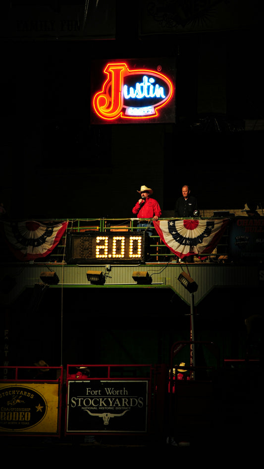 Two men stand near a digital clock at a rodeo.