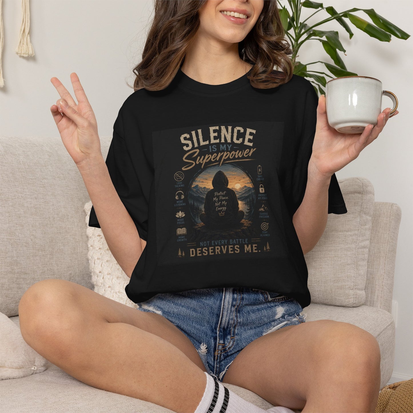 Black t-shirt featuring "Silence Is My Superpower" graphic, model sitting with a cup, casual style, peace sign gesture.