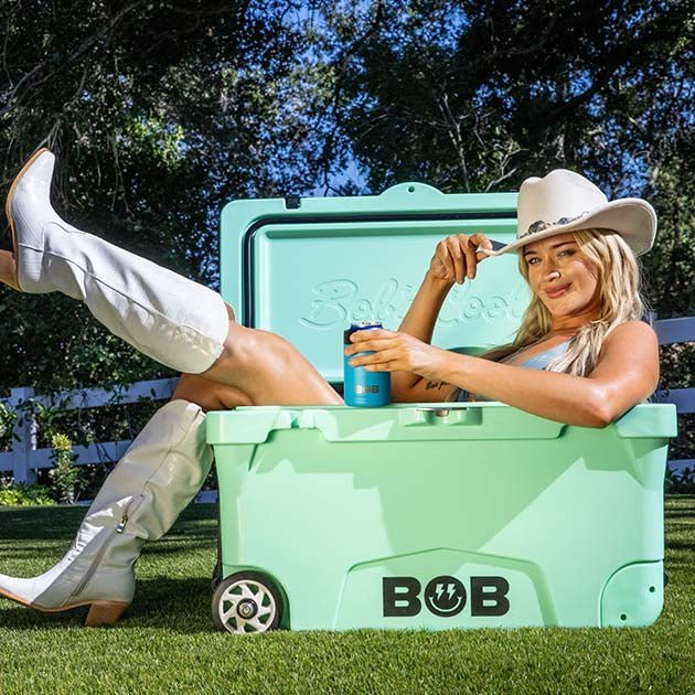 75QT "BFF" Hard Cooler in mint green with a woman in a cowboy hat and boots lounging on it outdoors.